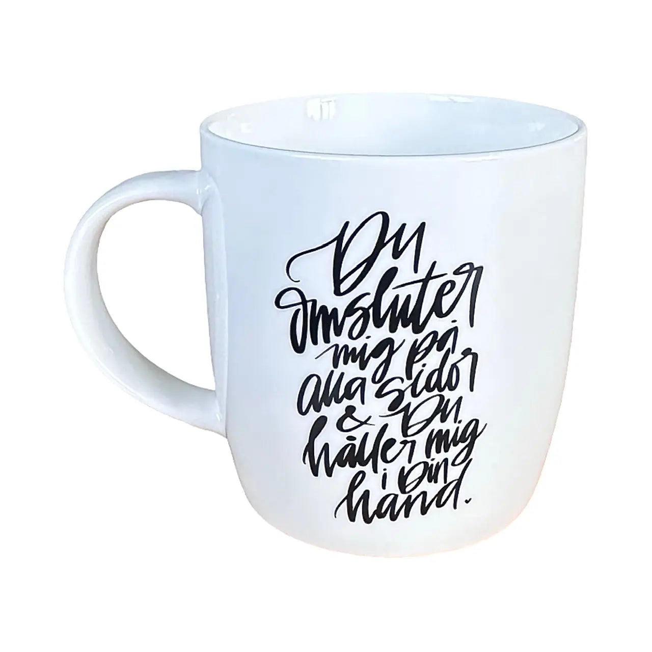 En stilren, naturvit mugg med svart text ”Du omsluter mig på alla sidor & Du håller mig i Din hand.”